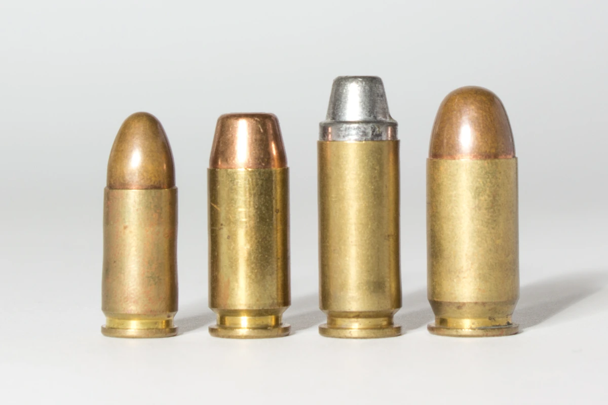 Four pistol cartridges standing upright side by side on a white ground. From the left: a slim round-nosed 9mm, a shorter flat-nosed .40 S&W of larger diameter, a distinctly taller 10mm Auto with a grey truncated-cone bullet, and a wide .45 ACP with a rounded jacketed bullet.