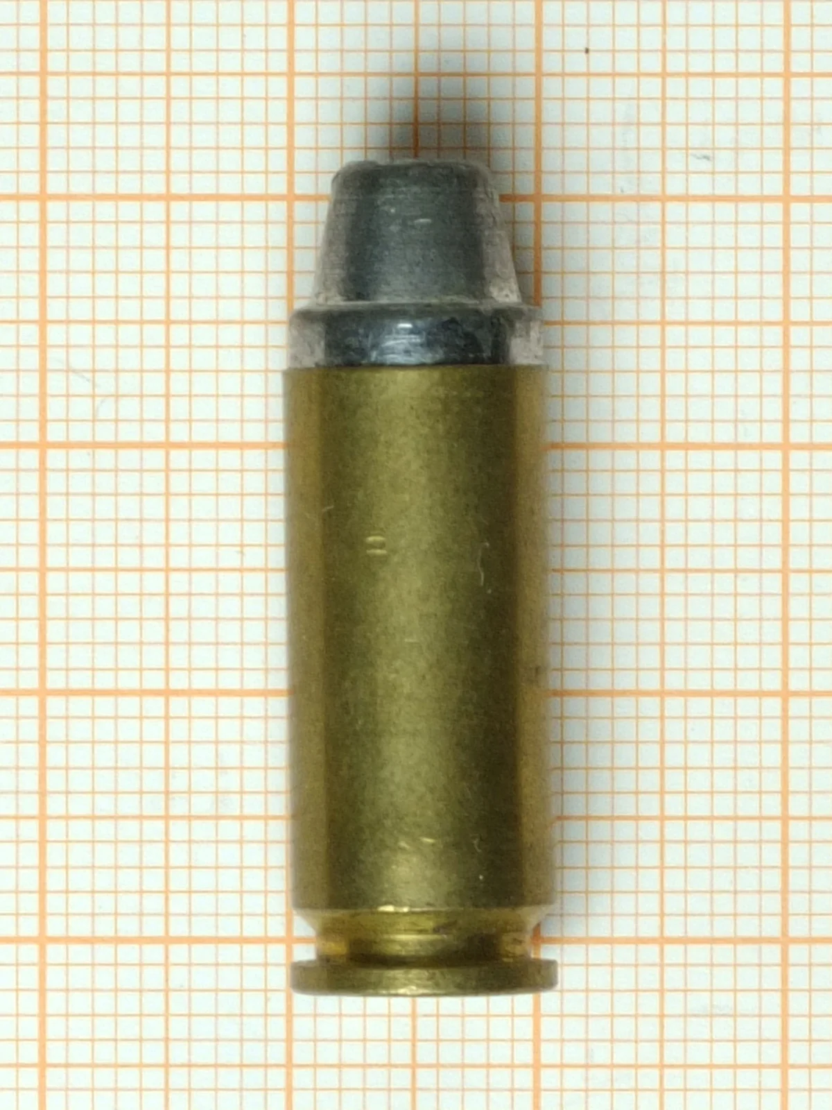 A single 10mm Auto cartridge lying on orange graph paper. The brass case is straight-sided with a wide extractor groove above the rim, and carries a grey truncated-cone lead bullet whose flat nose is about two thirds the width of the case.