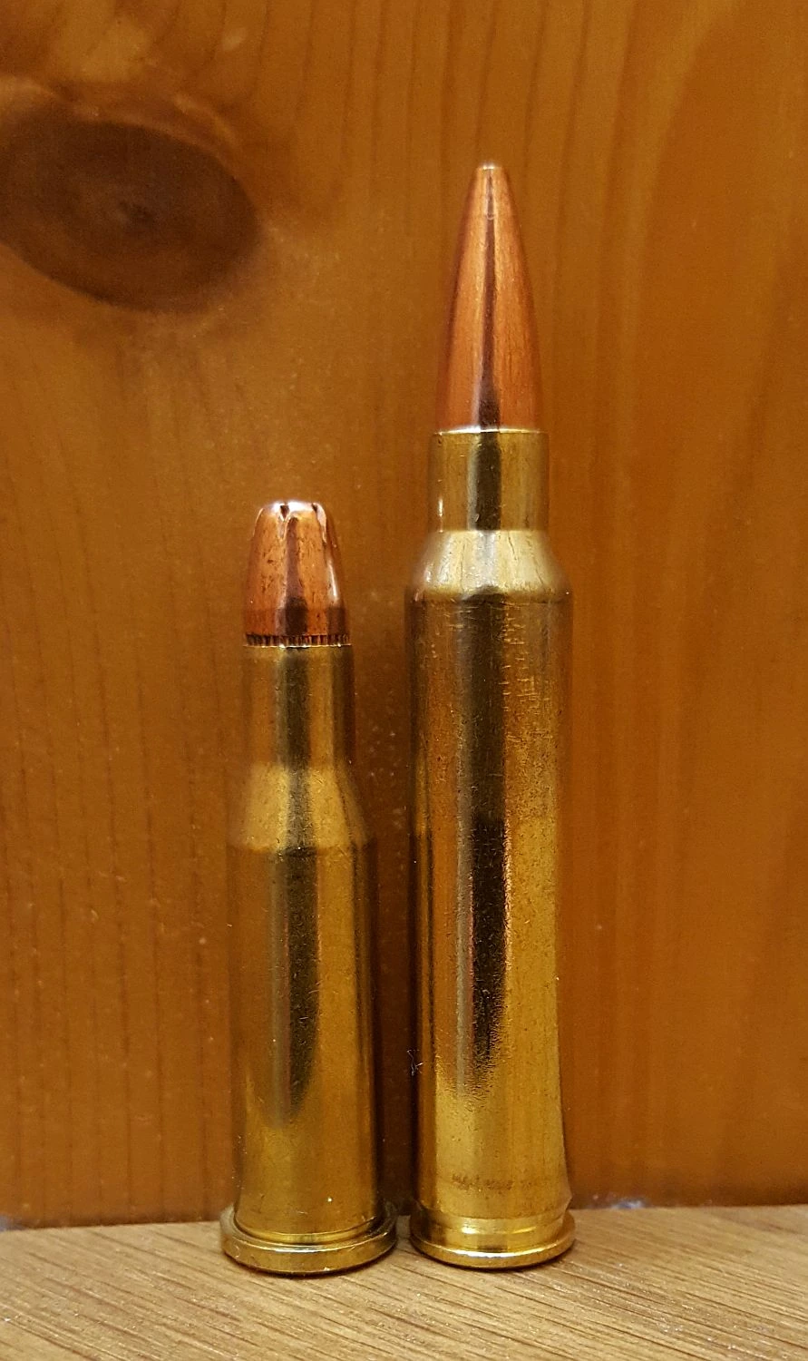 Two rimmed, bottlenecked rifle cartridges standing upright against a wooden background: a shorter one with a hollow-point bullet on the left, a taller one with a sharp spitzer bullet on the right.
