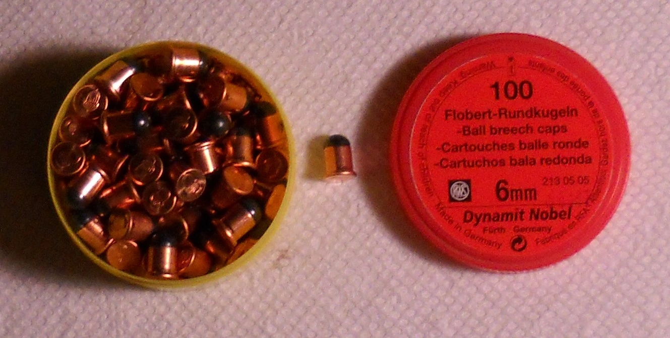 A round tin open to show dozens of tiny cartridges with round ball bullets, one loose cartridge beside it, and a red lid printed 'Flobert-Rundkugeln - Ball breech caps - 6mm - Dynamit Nobel'.