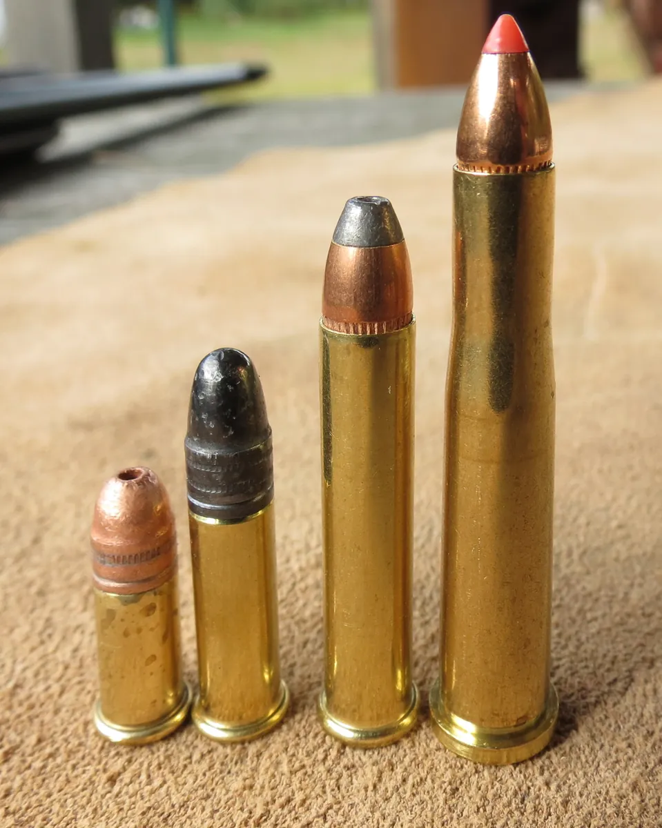 Four small brass cartridges standing on a sand-coloured surface, rising in height from left to right. The two on the left are short and plain-walled with a flat rim; the third is longer with a lead-tipped jacketed bullet; the tallest on the right is a bottlenecked case with a red-tipped copper bullet.
