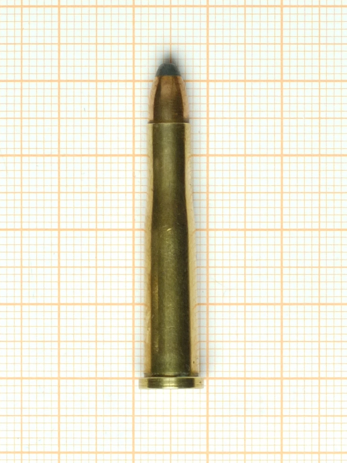 A single .22 Hornet cartridge lying on orange millimetre graph paper. The brass case is long, slim and noticeably tapered, ending in a short shoulder and a narrow neck, and carries a copper-jacketed bullet with an exposed grey lead tip. A thin rim projects at the base.
