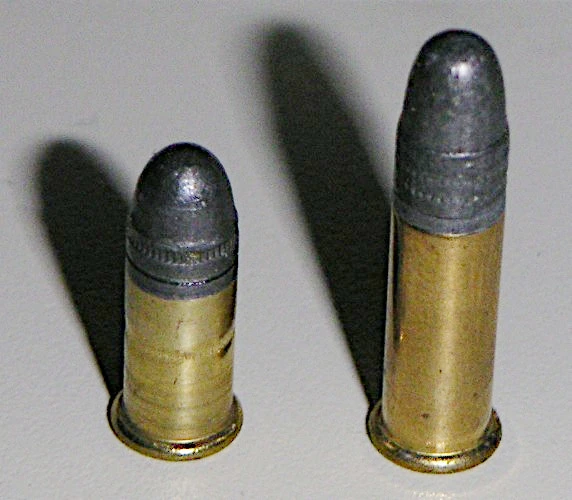 Two rimfire cartridges standing upright side by side on a pale ground: a shorter round-nose cartridge on the left and a taller one on the right.
