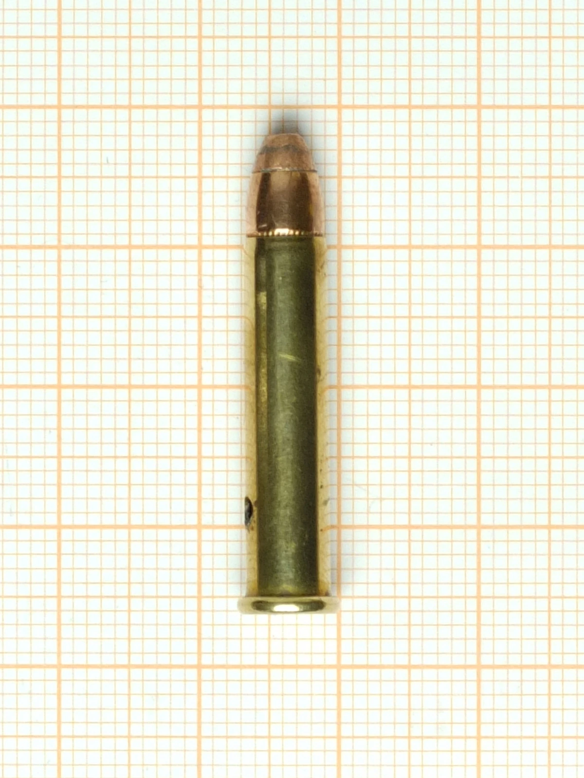 A single .22 Winchester Magnum Rimfire cartridge lying on orange graph paper, bullet uppermost. The bullet is copper-jacketed with a flattened nose, the brass case is straight-sided, and a thin rim projects at the base.