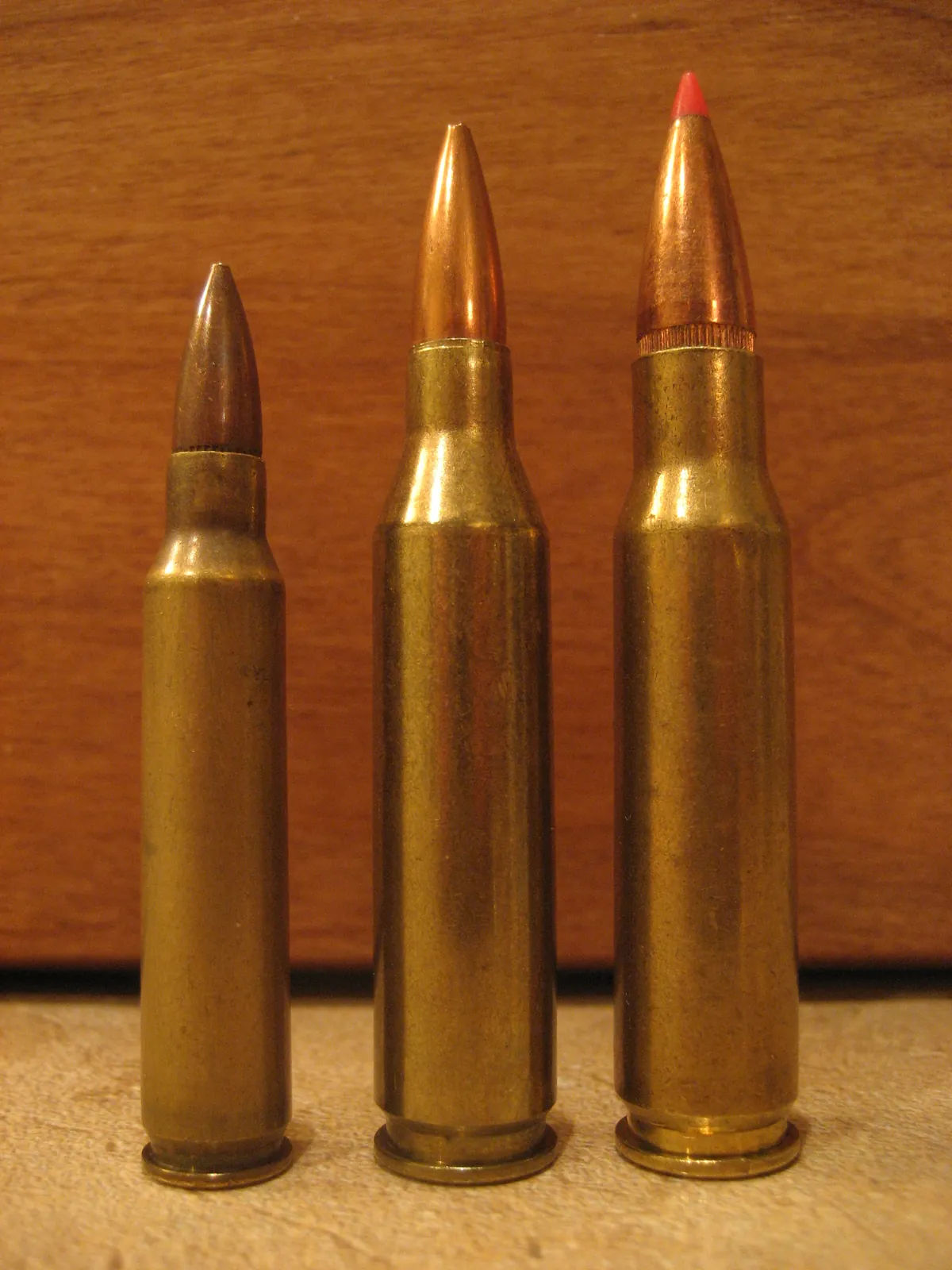Three bottlenecked brass rifle cartridges standing upright against a wooden panel. The left one is smallest, with a plain copper spitzer bullet; the middle is taller with a long slender copper bullet; the right is the same height but visibly wider at the neck and carries a red polymer-tipped bullet.