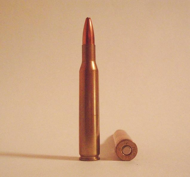 A single brass bottleneck rifle cartridge with a pointed copper jacketed bullet, standing beside its own base cap