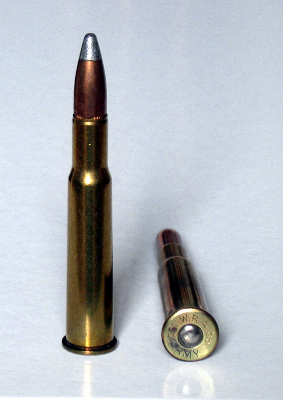 A .30-40 Krag rifle cartridge standing upright, a pointed soft-point bullet with an exposed lead tip seated in a long rimmed brass case, beside a second round lying on its side showing a W.R.A. Co. 30 Army headstamp.