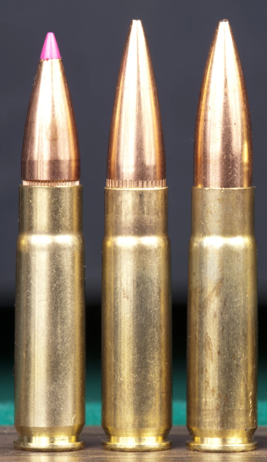 Three brass rifle cartridges standing upright against a dark background. The left has a pink polymer-tipped bullet, the centre a plain copper open-tip bullet, and the right a longer, more sharply pointed copper bullet seated deeper in a shorter visible case.