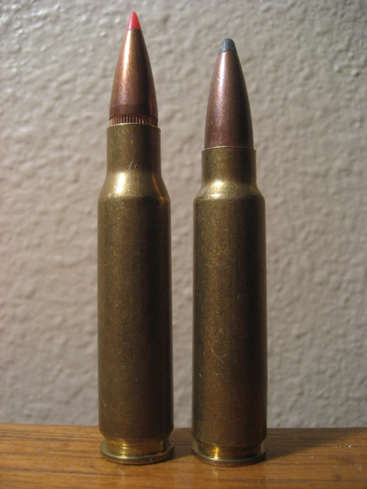 Two rimless bottleneck rifle cartridges standing upright side by side, the left one slightly taller with a red-tipped bullet, the right one with a plain lead-tipped bullet.