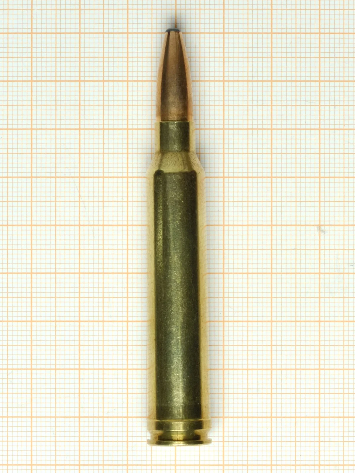 A single brass rifle cartridge with a belted case and a copper jacketed spitzer bullet, standing upright on graph paper.