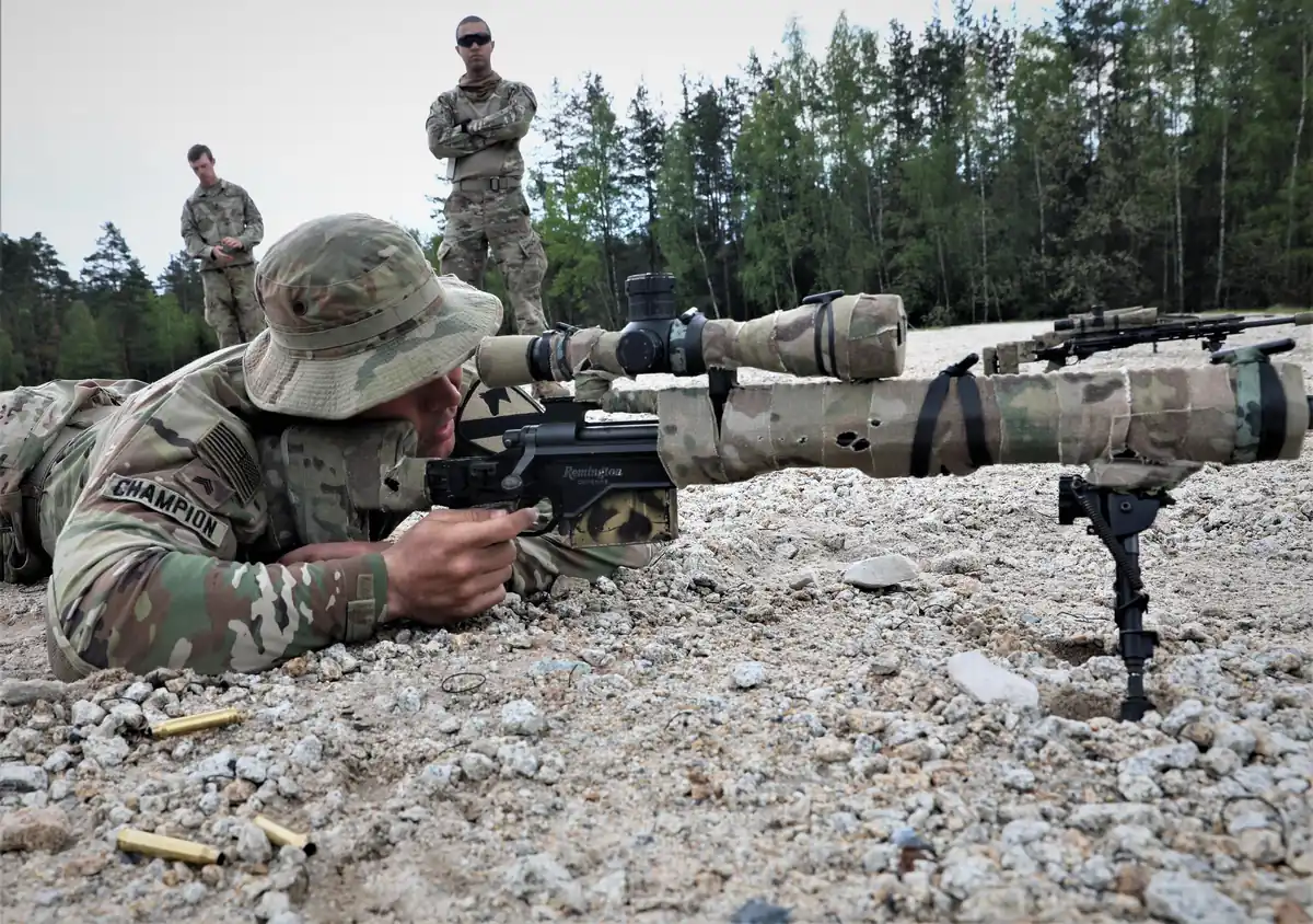 A soldier in camouflage and a boonie hat lies prone behind a heavy bolt-action rifle on a bipod, aiming down a large telescopic sight on a gravel range, with two spent brass cases on the ground and two soldiers standing watching behind him.