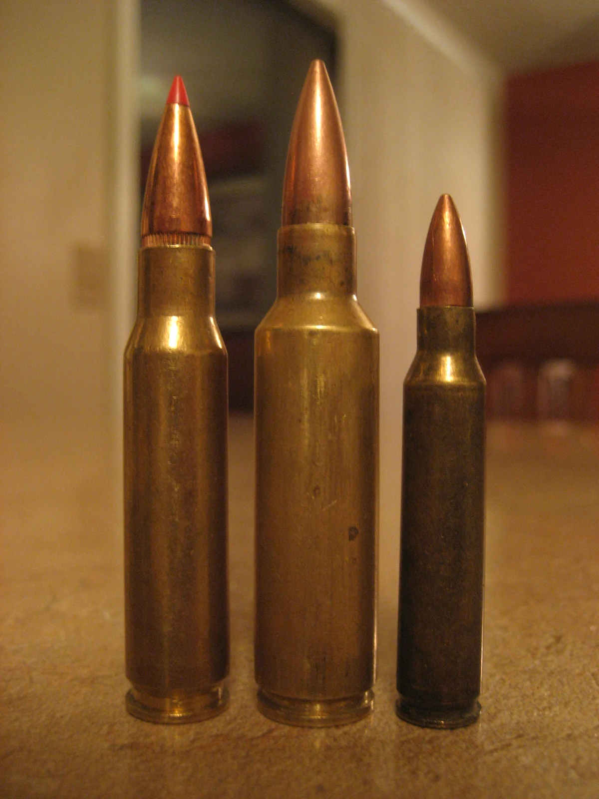 Three brass rifle cartridges standing upright: a shorter, wide-bodied round with a plain copper spitzer bullet in the centre, a taller round with a red polymer bullet tip to its left, and a smaller round with a dark case on the right.