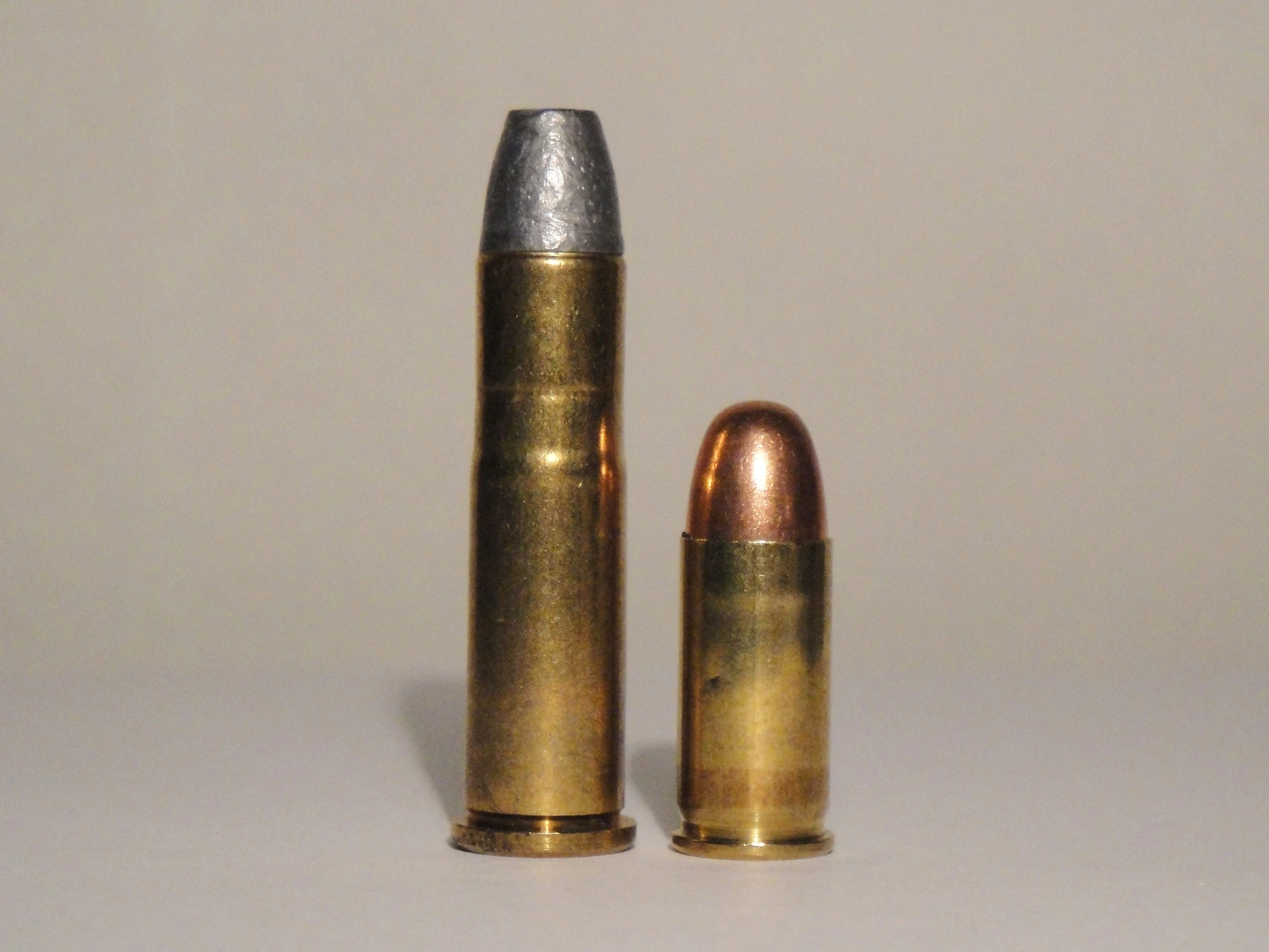 A tall, bottlenecked rifle-length cartridge with a flat, lead bullet standing beside a shorter, straight-walled pistol cartridge with a rounded copper-jacketed bullet.
