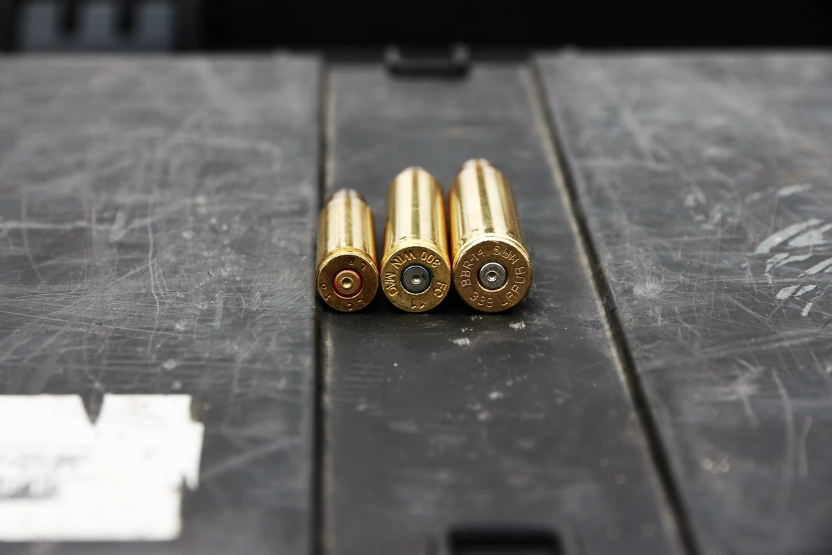 Three brass cartridge cases standing base-up on a scarred metal ammunition can, headstamps facing the camera and legible on the two right-hand rounds.