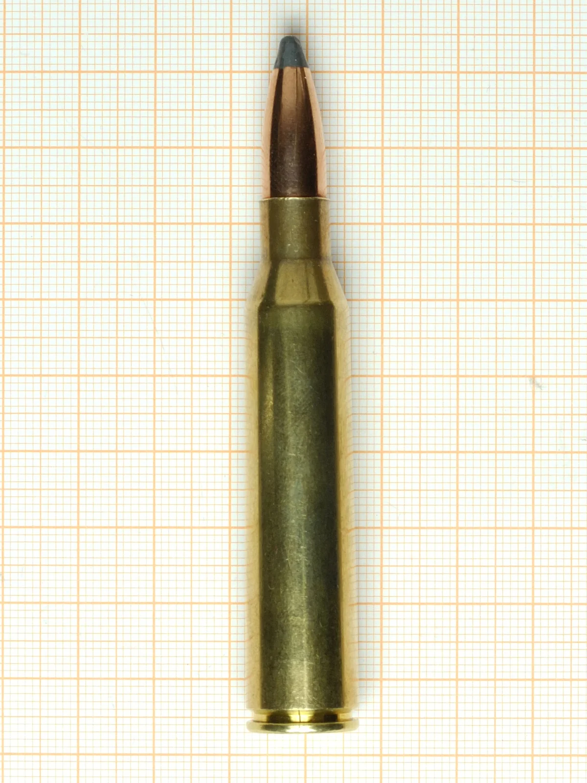A single brass rifle cartridge with a long copper jacketed bullet tipped in dark green, standing upright on graph paper.