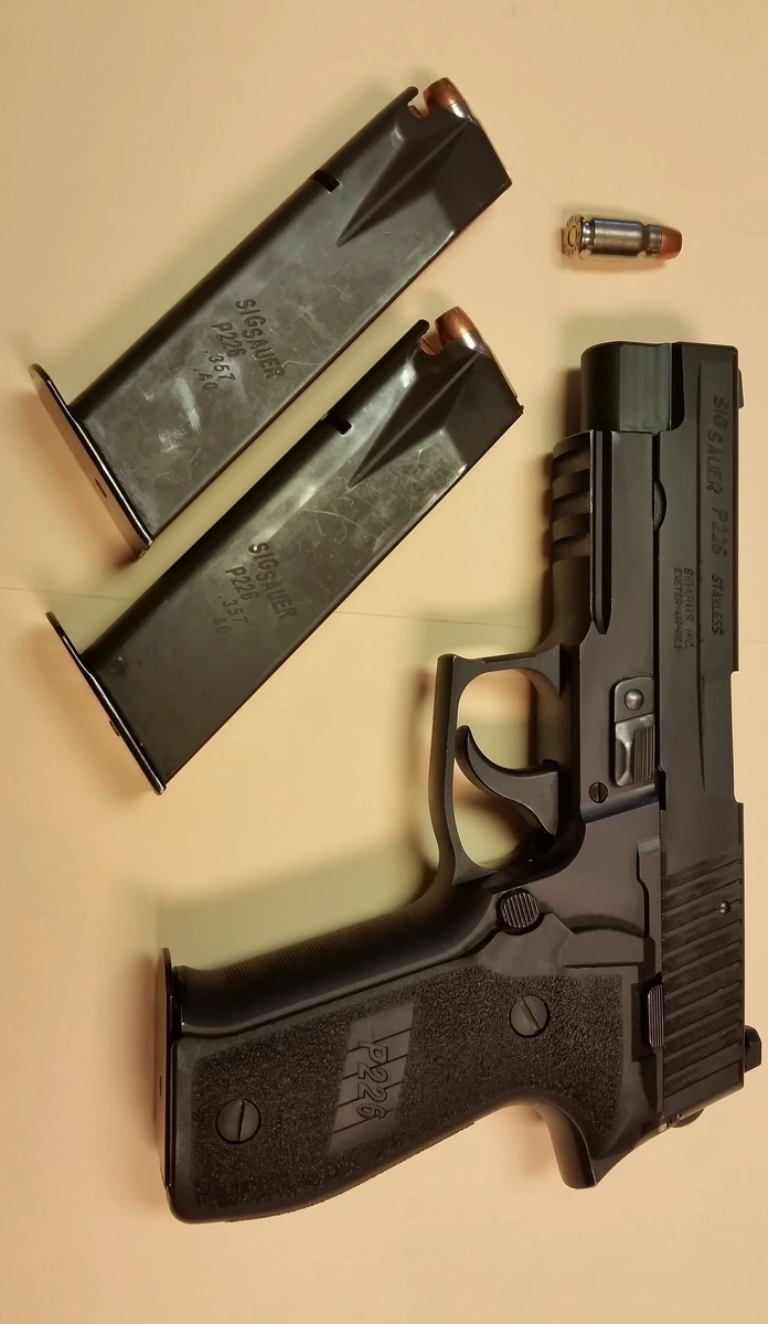 A black semi-automatic pistol lying on its side on a tan surface, beside two loaded box magazines marked SIGSAUER P226 .357 and a single loose cartridge with a jacketed hollow-point bullet.