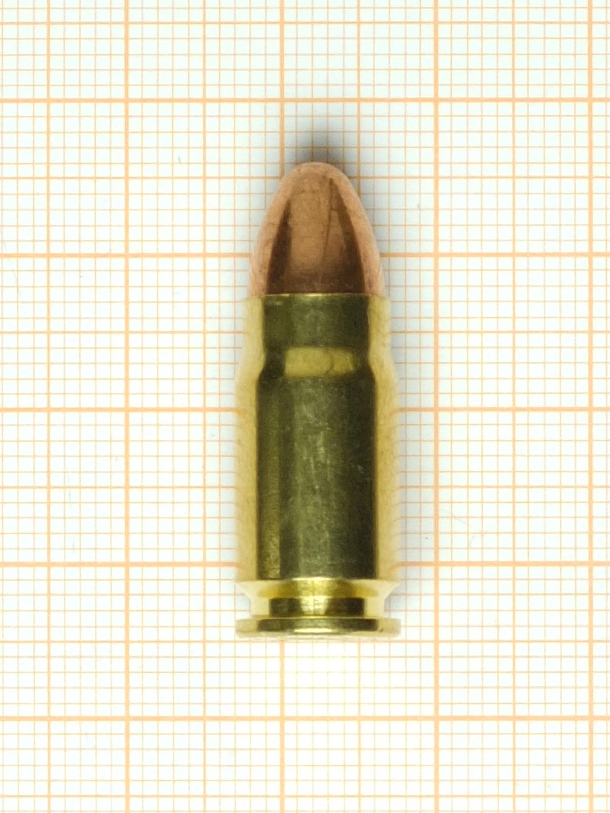 A single brass pistol cartridge with a bottlenecked case and a copper jacketed round-nose bullet, standing upright on graph paper.