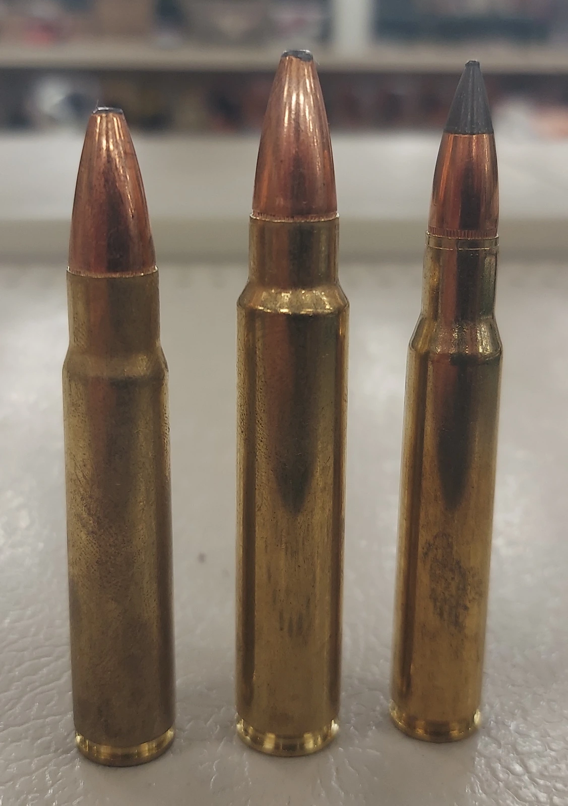 Three brass rifle cartridges of similar length standing in a row, the middle one topped by a wide copper bullet noticeably thicker than the other two.