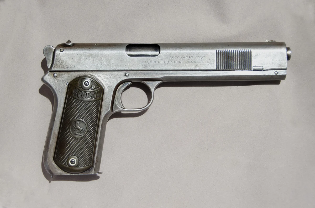 A worn nickel-finish semi-automatic pistol with chequered wooden grips bearing a Colt medallion, its slide marked 'AUTOMATIC COLT CALIBRE 38 RIMLESS SMOKELESS'.