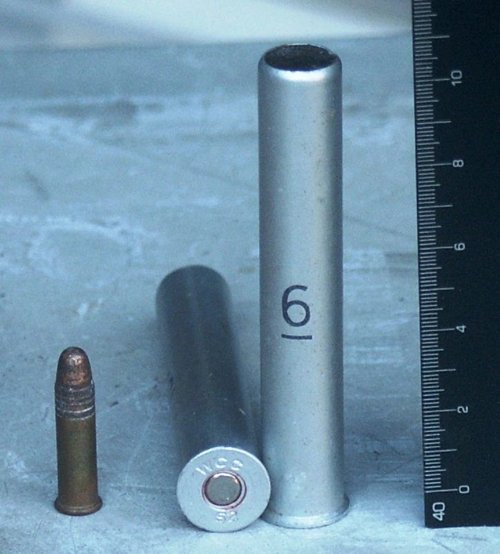 Two all-metal .410 cases beside a .22 Long Rifle cartridge, with a steel rule at the right. One .410 case stands upright showing its open mouth; the other lies on its side showing the headstamp W C C 52. The .410 cases are about three times the diameter of the rimfire round.