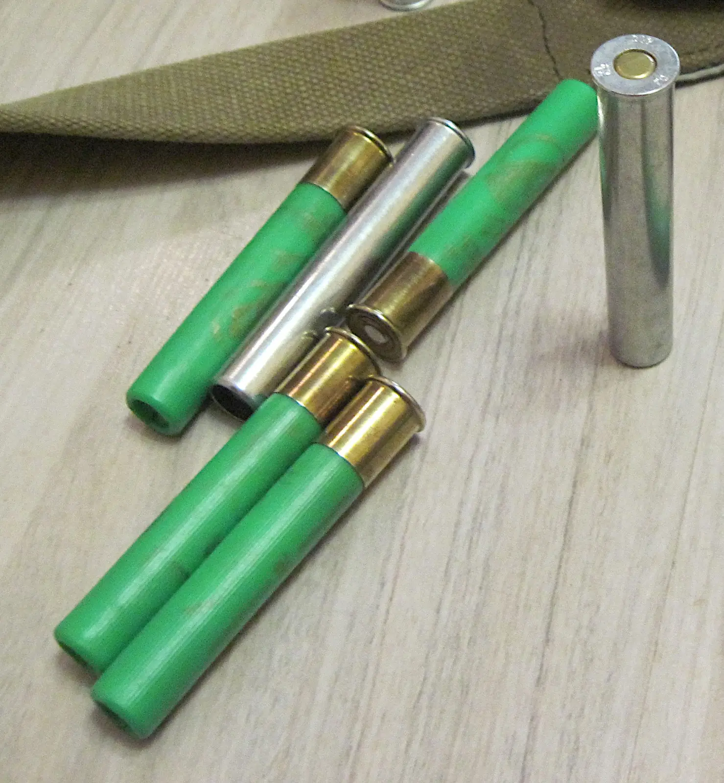 Six empty .410 bore cases lying on a pale wooden surface: four with green plastic hulls and brass heads, and two plain nickel-coloured all-metal cases, one of them standing upright showing its primer and headstamp.