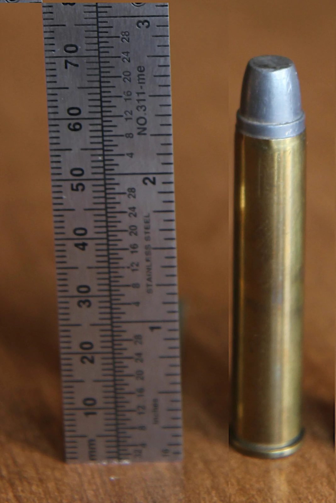 A .444 Marlin rifle cartridge standing upright beside a steel ruler, showing a flat-tipped soft-lead bullet with a crimped gas check seated in a long straight brass case.