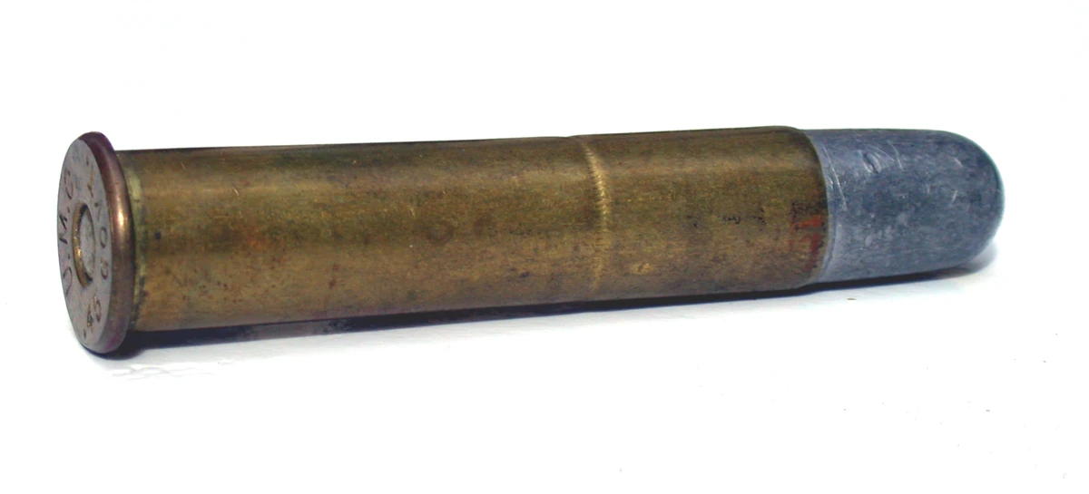 A single old .45-70 cartridge lying diagonally on a white ground. The brass case is dark and tarnished, with a copper-coloured rim carrying a worn headstamp, and it is loaded with a plain grey lead round-nosed bullet that shows no jacket.