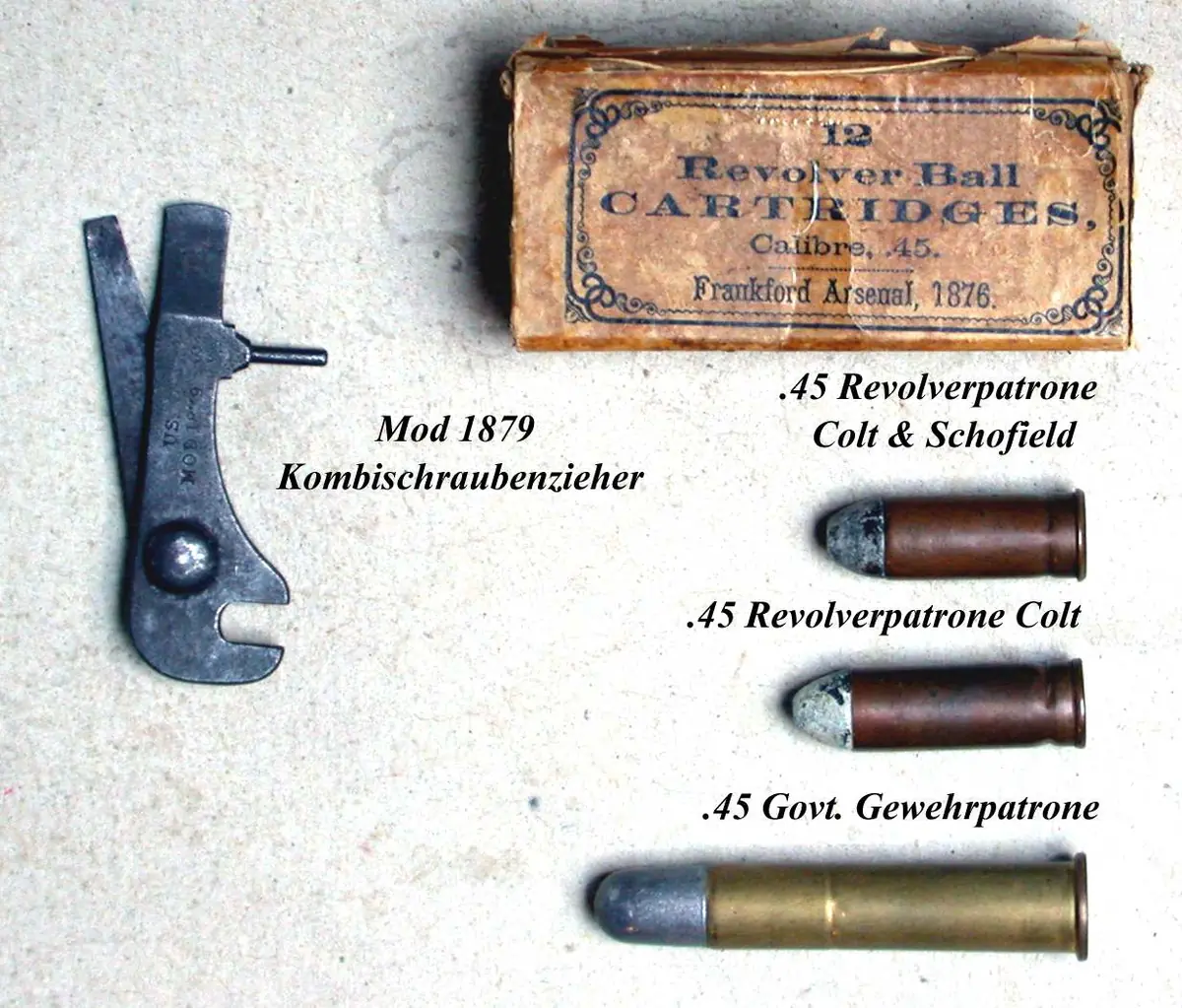 A worn paper cartridge box printed "12 Revolver Ball Cartridges, Calibre .45, Frankford Arsenal", above three loose lead-bulleted cartridges and a small steel combination tool, each labelled in German.