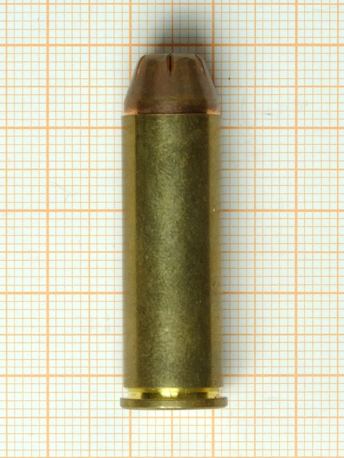 A single straight-walled brass revolver cartridge with a copper-jacketed hollow-point bullet and a narrow rim, standing upright on orange millimetre graph paper.