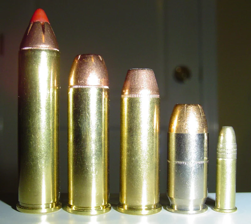 Five brass cartridges standing in a row, decreasing in length from left to right; the tallest, on the left, has a red polymer-tipped bullet.