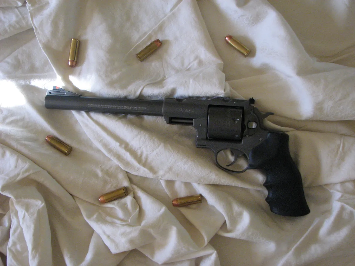 A dark stainless double-action revolver with a long barrel and a rubber grip lying on a white sheet, with several brass cartridges scattered around it.
