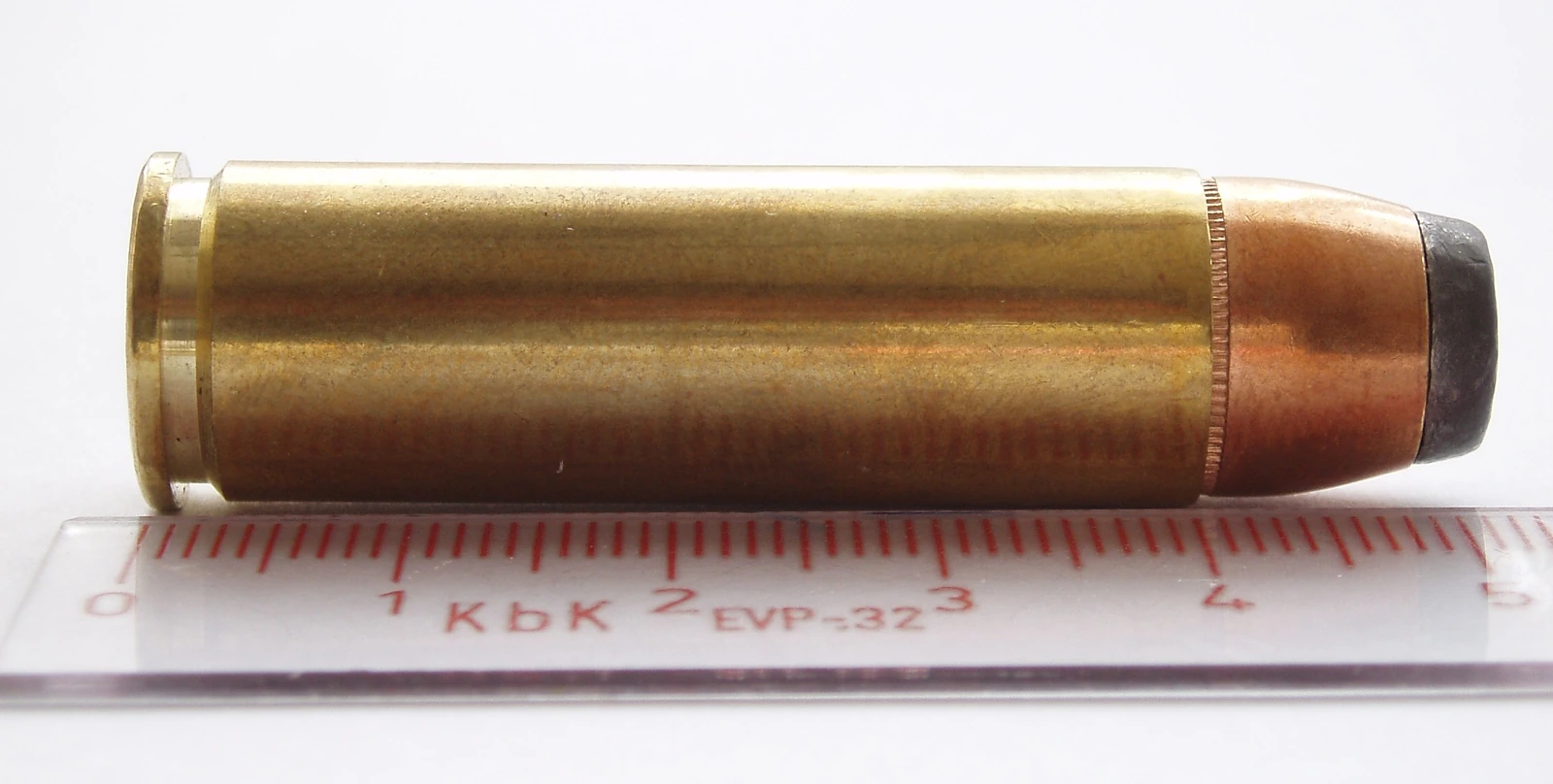 A single long, wide brass revolver cartridge with a soft-point bullet lying on its side above a transparent ruler marked in centimetres.