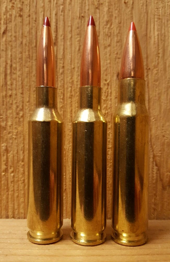 Three brass rifle cartridges standing upright against a pine board. All three carry copper jacketed bullets with red polymer tips. The left two are slender and near-identical in height; the right one is visibly shorter in the body and wider at the shoulder.