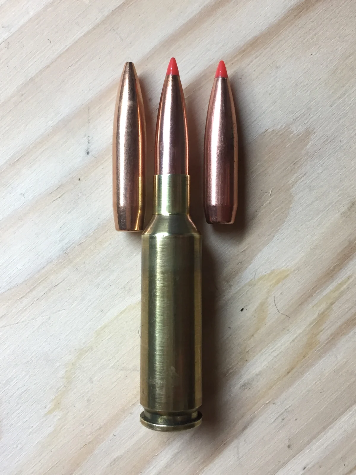 A slender bottlenecked brass rifle cartridge with a red-tipped bullet standing upright on light wood, flanked above by two long loose bullets.