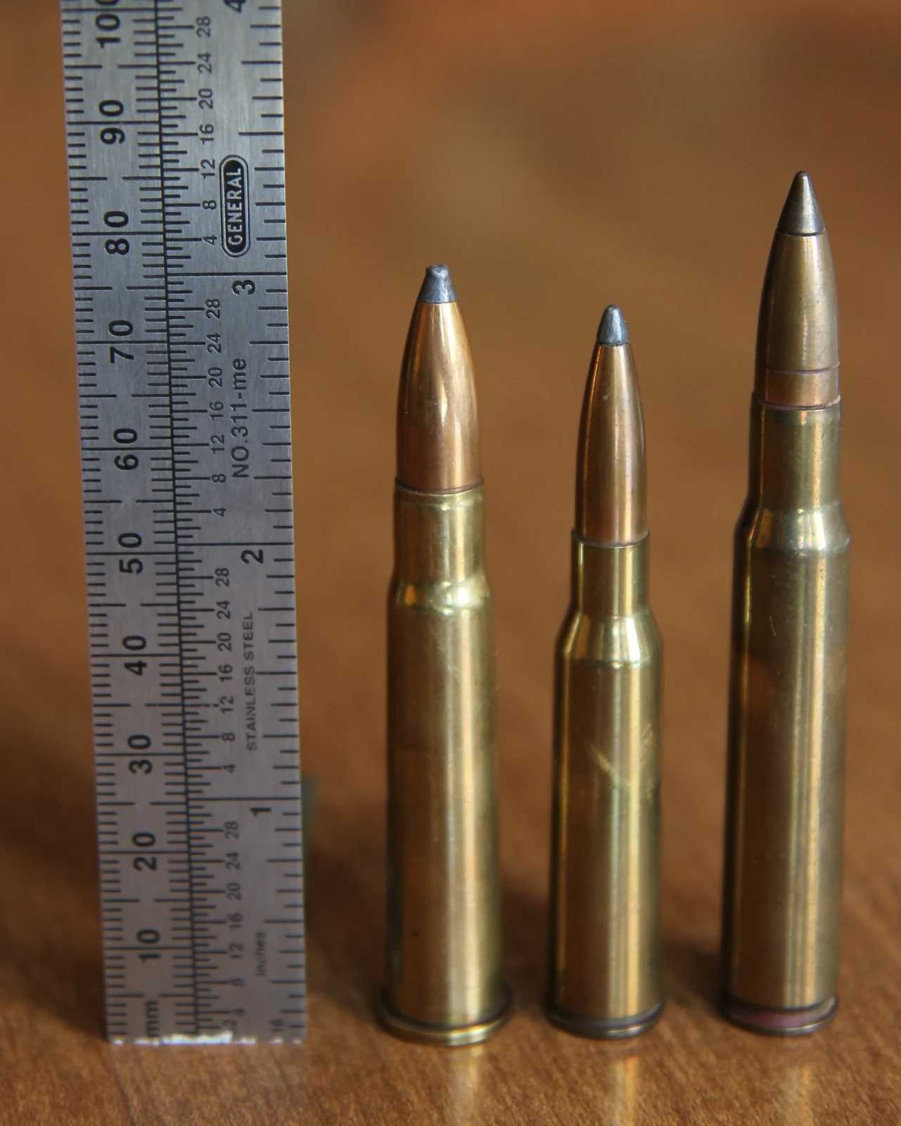 Three bottlenecked brass rifle cartridges standing on a wooden table beside a steel rule marked in millimetres and inches. The middle one is markedly slimmer than the other two and carries a lead-tipped copper bullet; the left has a wider body and a blunter bullet; the right is the tallest, with a jacketed bullet showing a cannelure.