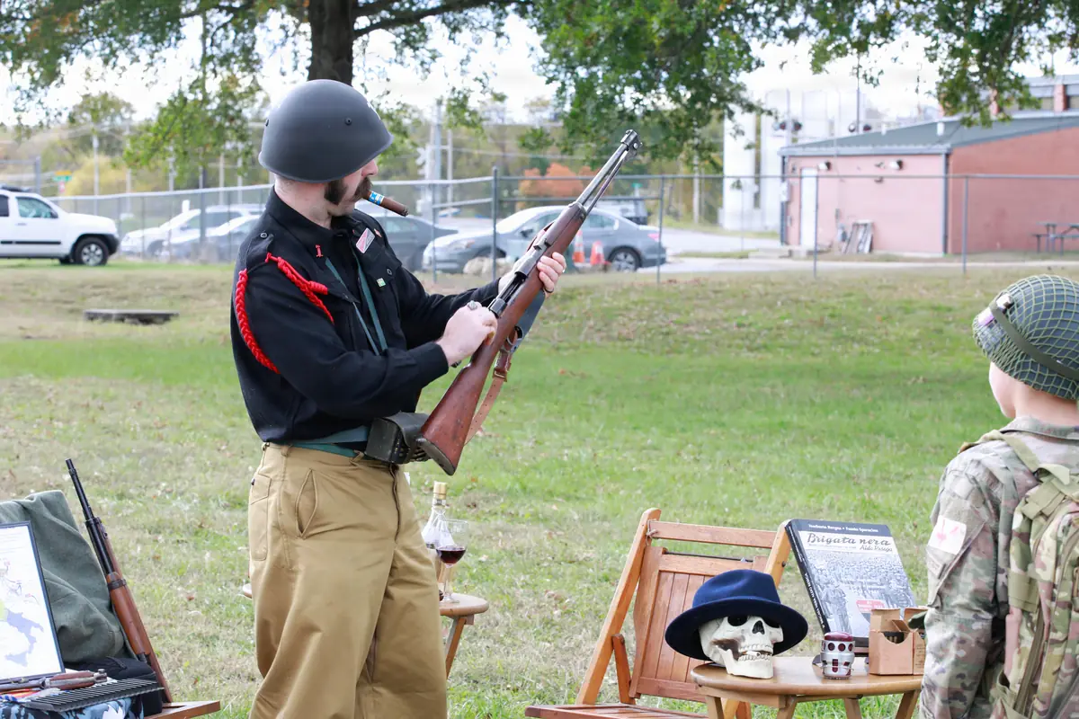A military re-enactor in period dress demonstrating a Carcano rifle to a visitor outdoors