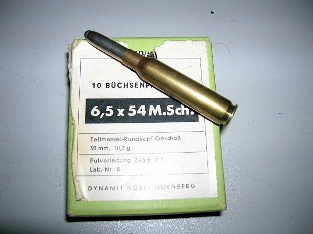 A single 6.5x54mm Mannlicher-Schoenauer rifle cartridge standing beside a period box printed 6,5 x 54 M.Sch. in German