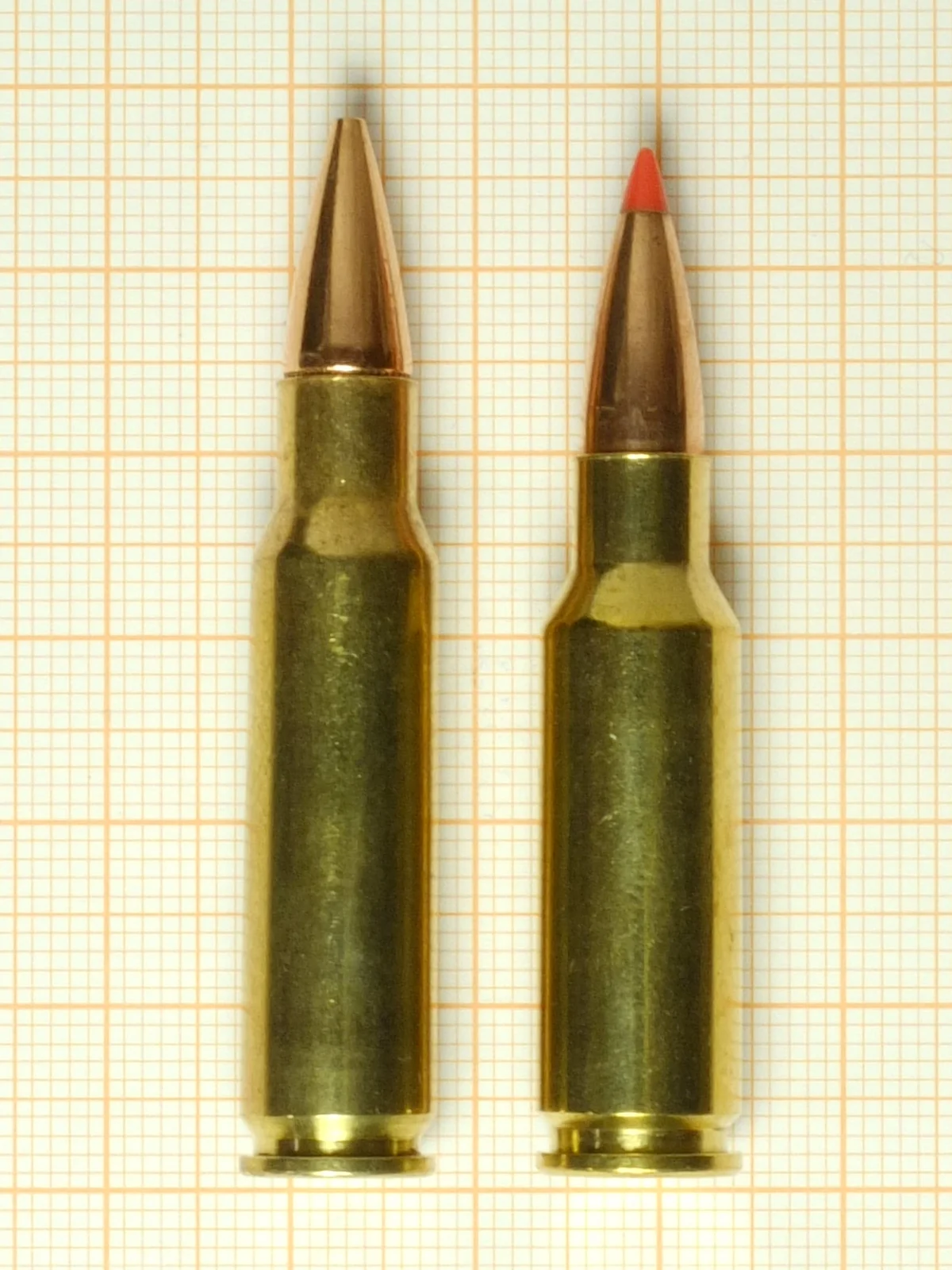 Two rifle cartridges standing side by side on graph paper, the left one shorter with a copper-and-red bullet tip, the right one taller with a plain copper tip.