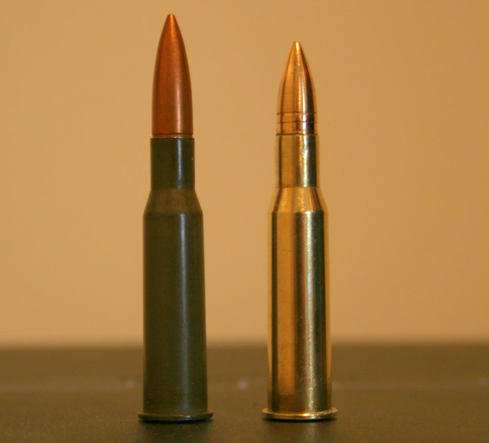 Two rimmed rifle cartridges standing upright side by side: one with a lacquered-steel case and copper-jacketed bullet on the left, one with a plain brass case and a bullet showing cannelure marks on the right.