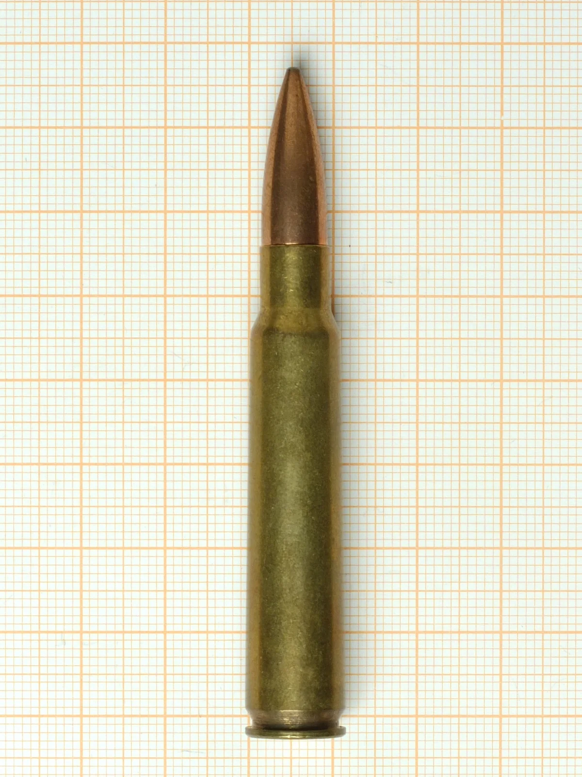 A 7.7×58mm Arisaka rifle cartridge lying on graph paper, a pointed copper-jacketed spitzer bullet seated in a long brass bottlenecked case.