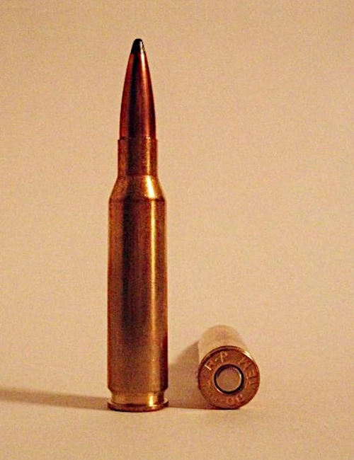 A rifle cartridge standing upright beside its own spent case laid on its side, showing the headstamp, against a plain tan background. The cartridge has a brass bottlenecked case and a copper jacketed bullet with a dark tip.