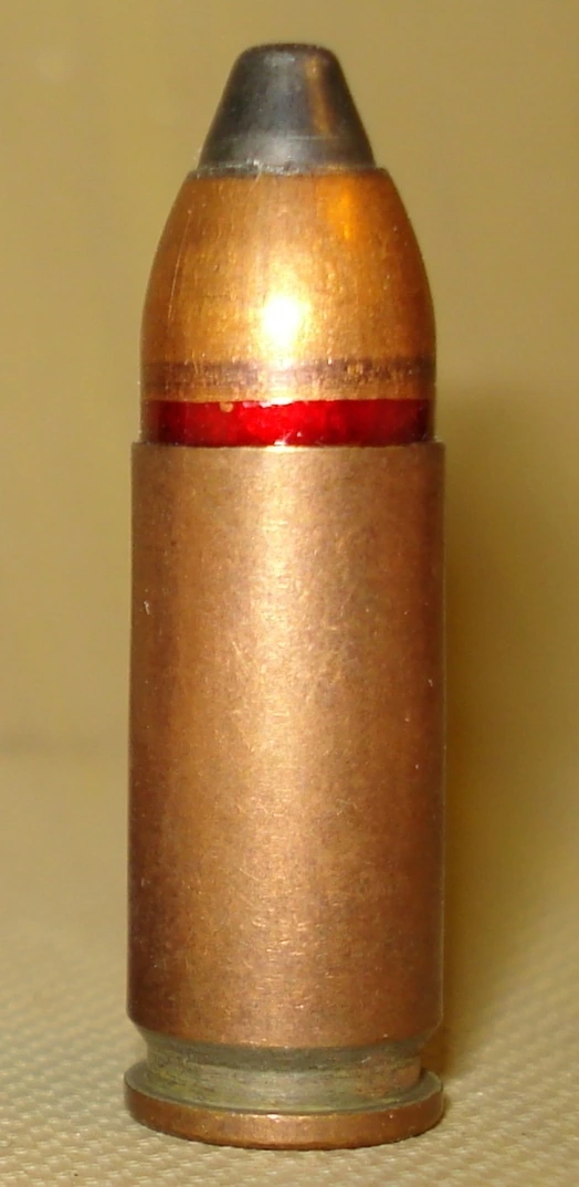 An upright straight-walled pistol cartridge with a red sealant band at the case mouth and a bullet with a dark steel-coloured tip.