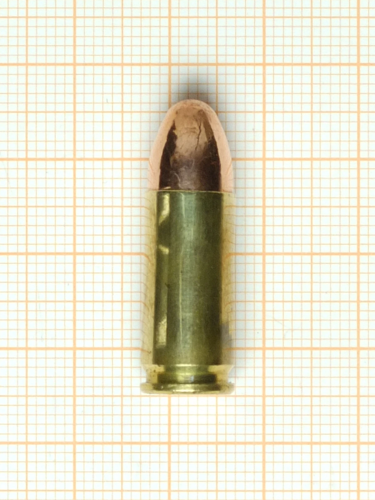 A single brass pistol cartridge with a round-nose copper jacketed bullet, standing upright on graph paper.