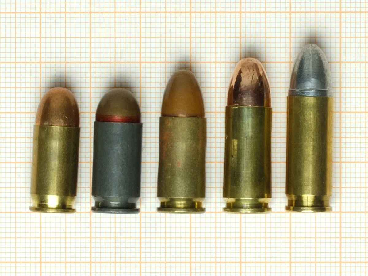 Five nine-millimetre pistol cartridges lying side by side on orange graph paper, rising in length from left to right. The second from the left has a dark grey steel case with a red sealant ring; the rest are brass. The rightmost is the longest and carries a lead-cored round-nosed bullet.