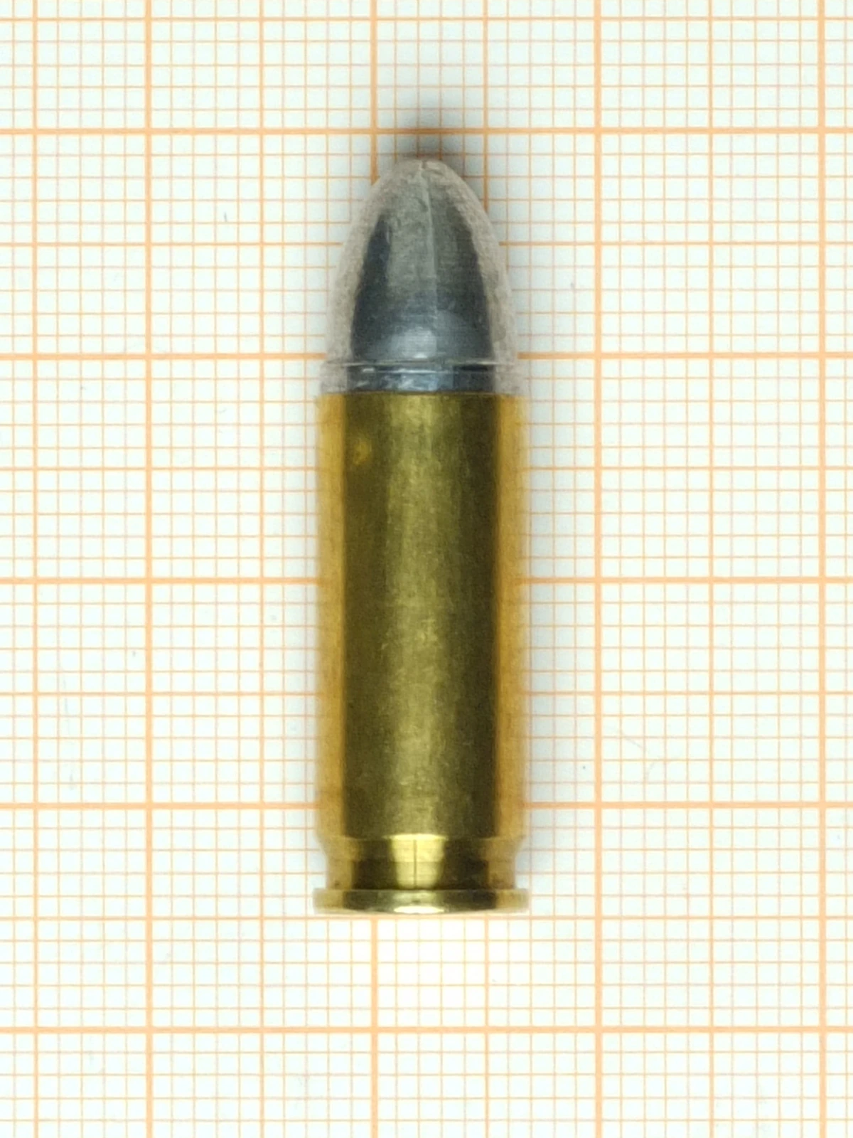 A single 9x23mm Steyr cartridge on orange millimetre graph paper. The brass case is straight-sided and about two and a half times as long as it is wide, and carries a round-nosed bullet whose grey lead core shows through a translucent jacket. The base is flat with no projecting rim.