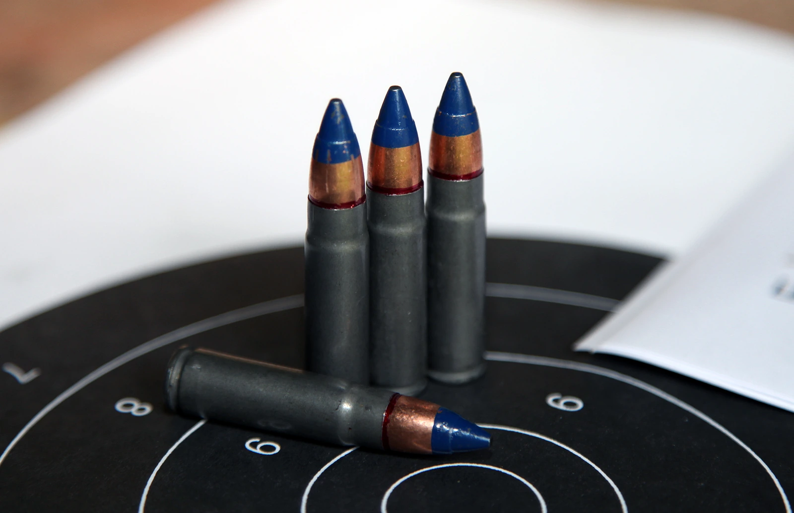 Four cartridges with dull grey lacquered steel cases on a black-and-white paper target. Three stand upright and one lies in front. Each carries a stubby copper-jacketed bullet whose forward half is painted bright blue, sealed at the case mouth with a red ring.