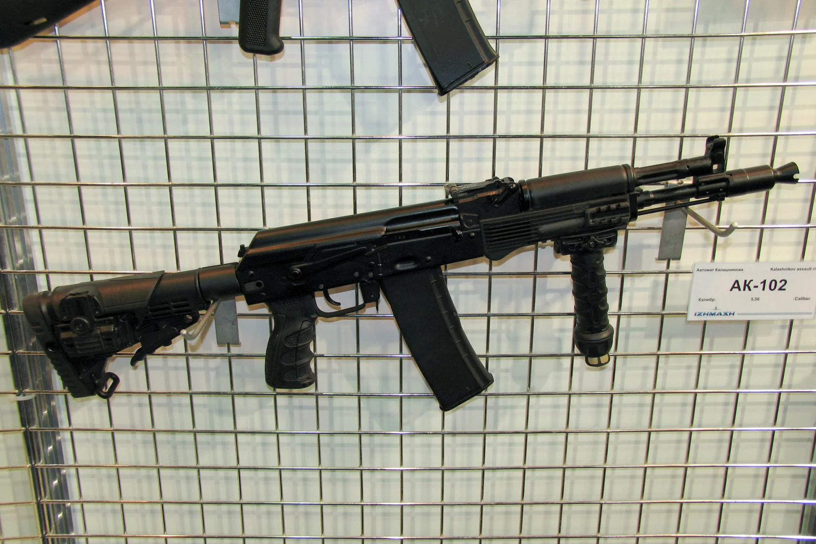 A black AK-102 carbine with a side-folding stock, a vertical foregrip and a curved magazine, displayed hanging on a wire panel at an arms exhibition