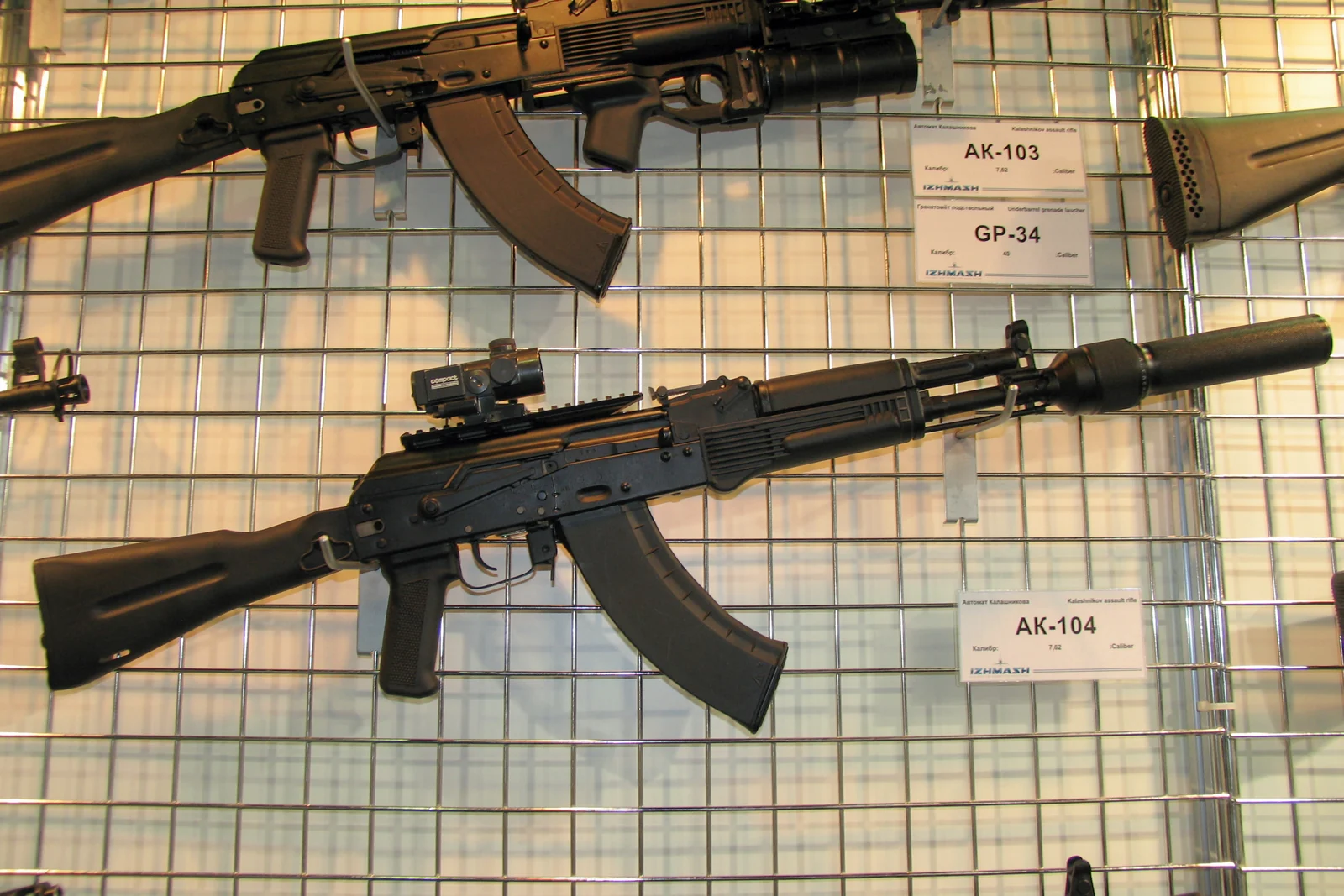 A black AK-104 carbine with a red dot sight mounted on a top rail and a suppressor attached to the barrel, displayed on a wire panel at an arms exhibition