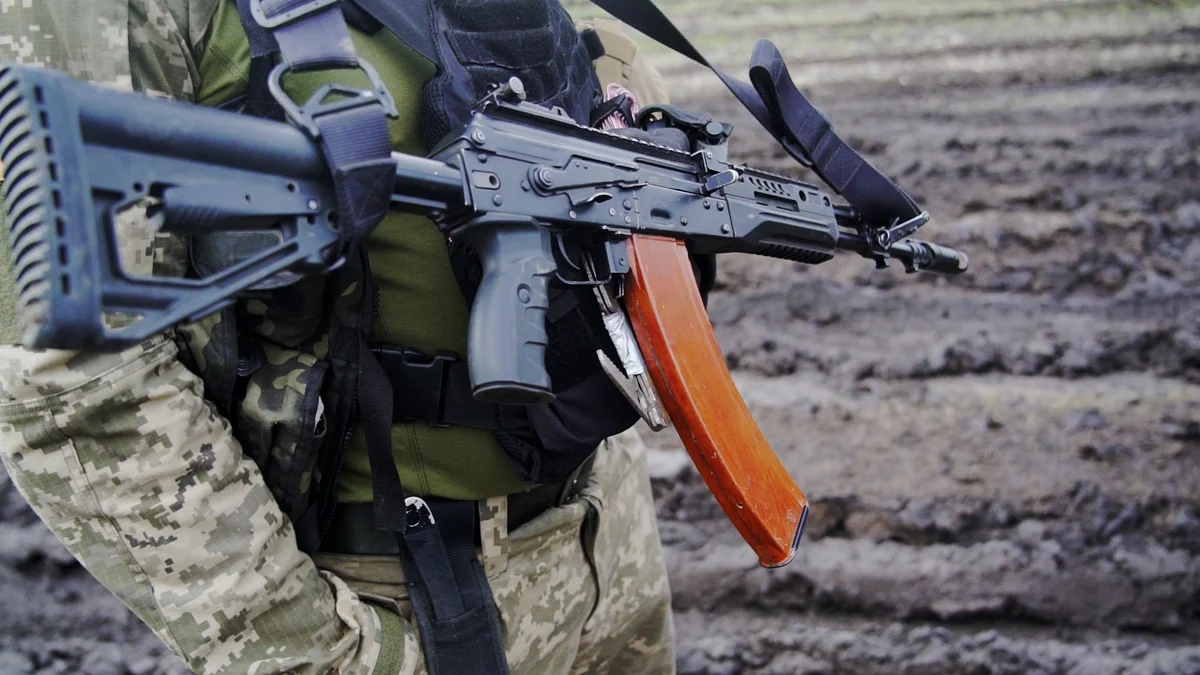 A soldier in field gear holding a black rifle with a rust-coloured curved magazine and a folding stock, slung across his chest