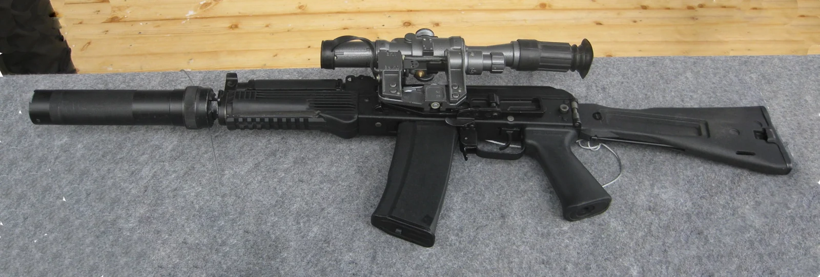 A black Kalashnikov-pattern carbine with a quick-detachable cylindrical suppressor and a large day/night optical sight mounted on a side rail, lying on a grey mat.